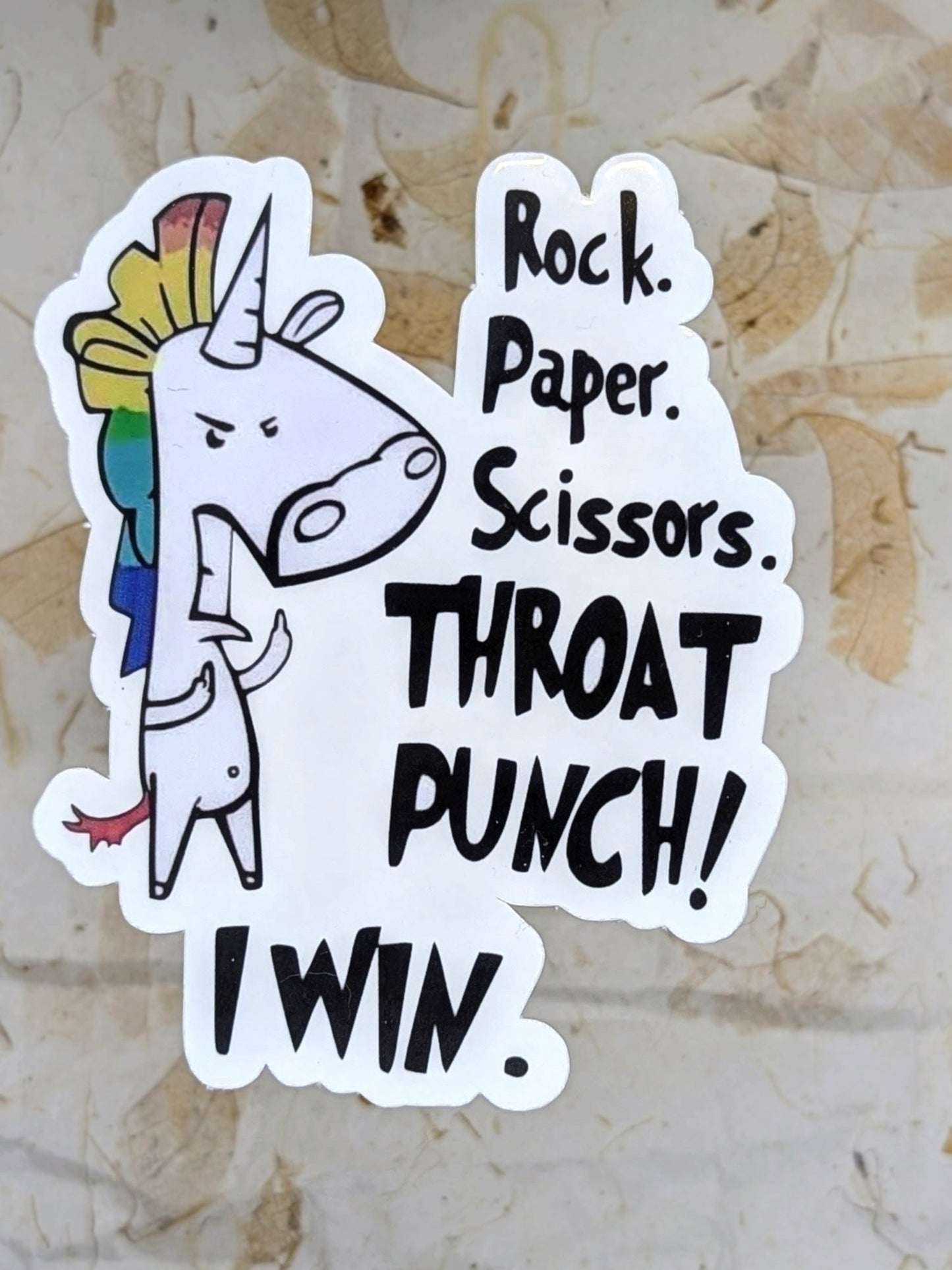 A die-cut sticker featuring a cartoon white unicorn with a rainbow mane and the text 'Rock. Paper.