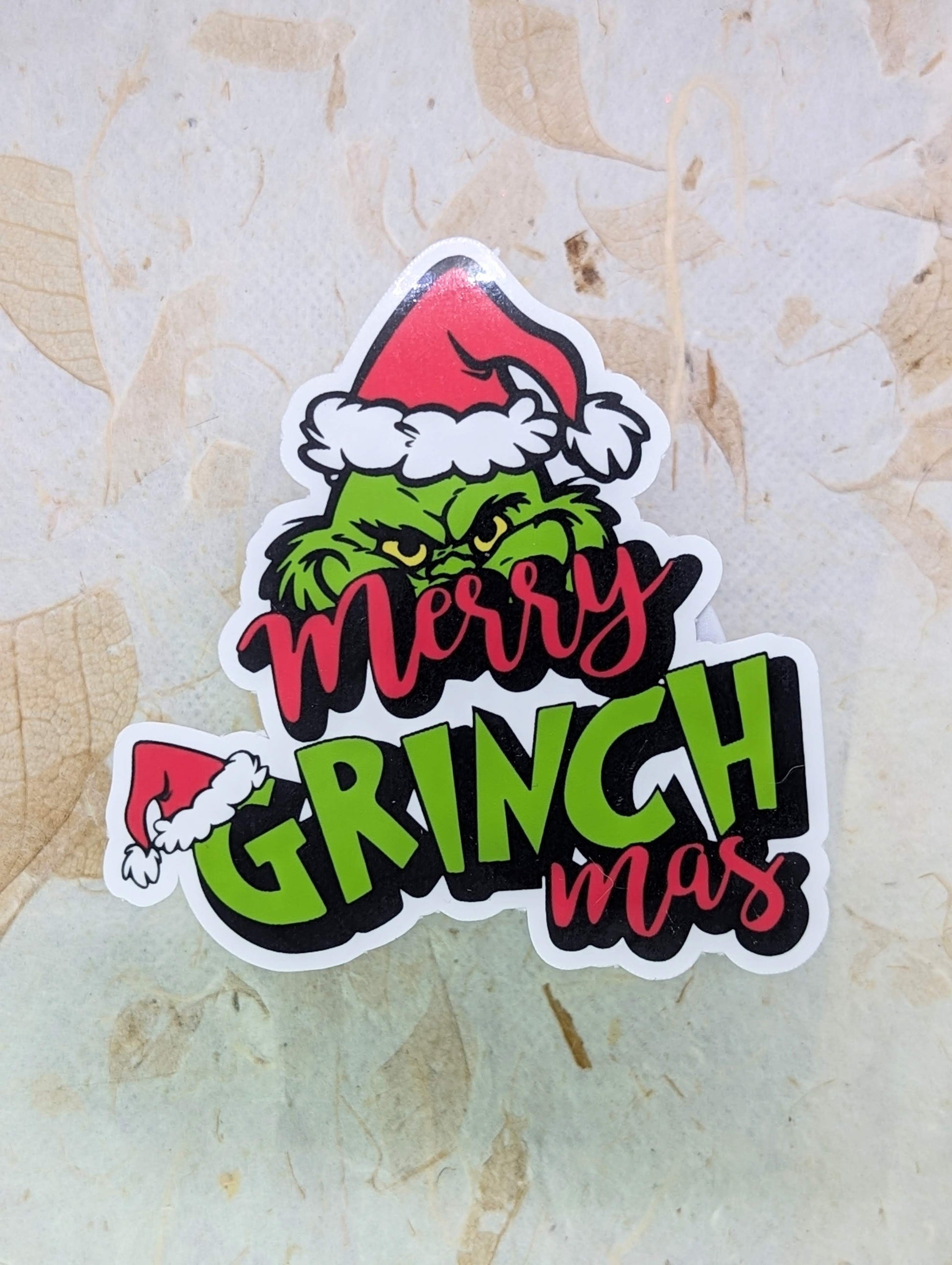 Sticker with Grinch design and 'Merry Grinchmas' text on a textured surface