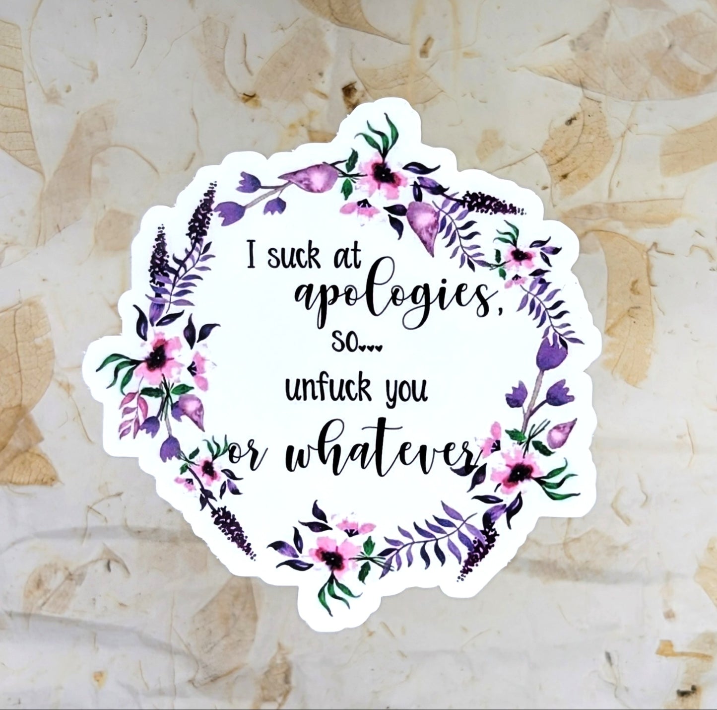 A die-cut sticker with a white background, featuring a watercolor floral wreath in purple, pink, and green, with black text that reads 'I suck at apologies, so.