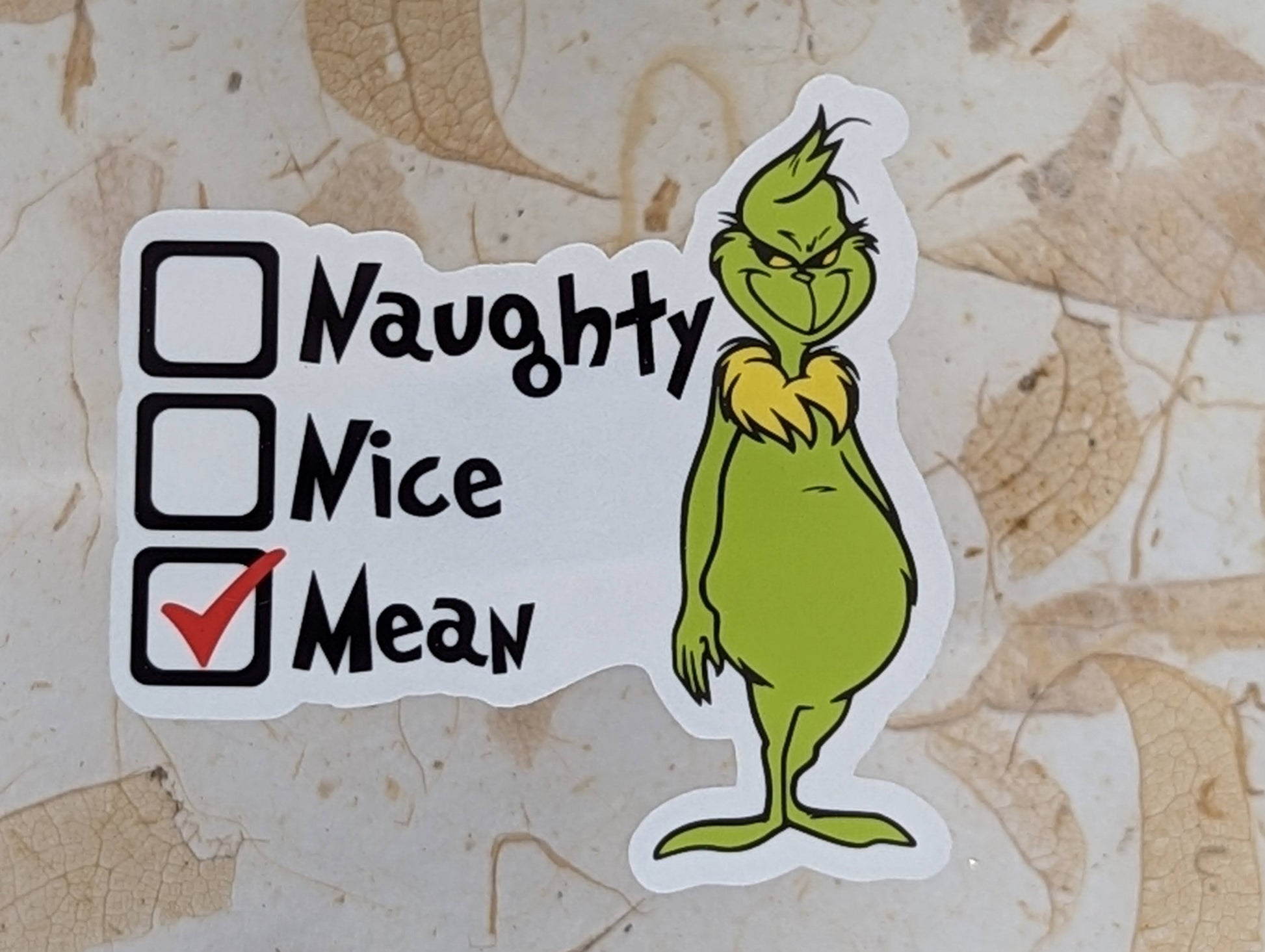 Sticker of a green character with checkboxes labeled 'Naughty', 'Nice', and 'Mean' on a textured beige background.