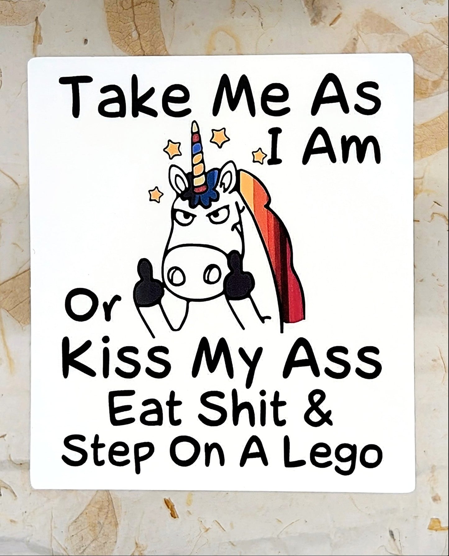A square white novelty sign with black hand-drawn text and a cartoon angry unicorn with a rainbow mane, placed on a light wood surface. The text reads: 'Take Me As I Am Or Kiss My Ass Eat Shit & Step On A Lego'.